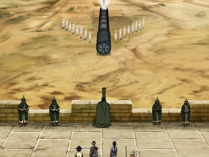 A group of characters watch a large mechanized drill advance toward a walled city in a desert landscape, from an animated TV show scene