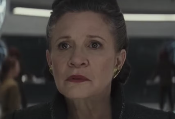 A person with an intense expression stands indoors, wearing a high-collared garment and gold earrings