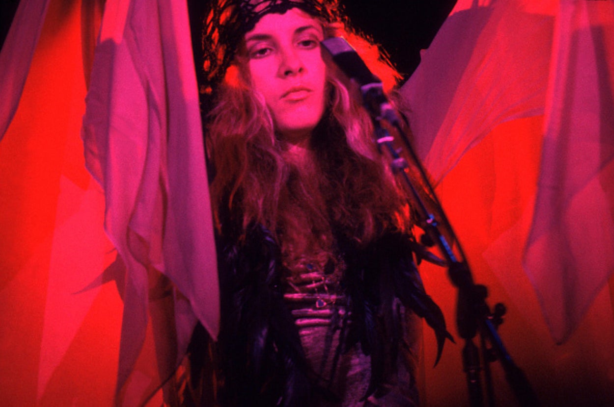 Person with long hair, wearing a feathered outfit and headband, stands at a microphone with flowing fabric as a backdrop