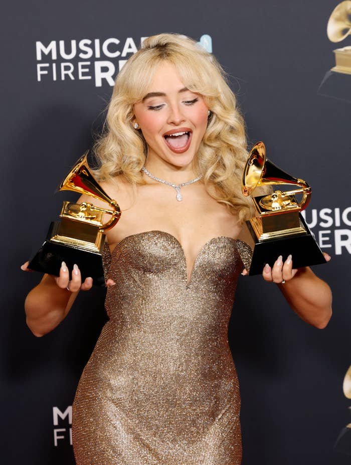 Person in a strapless sequin gown joyfully holds two Grammy awards at a music event