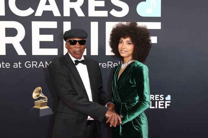 Two individuals on a red carpet, one wearing a tuxedo and cap, the other in a velvet evening gown, posing at a music relief event backdrop