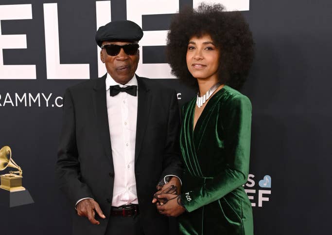Two individuals on a red carpet; one in a black suit and cap, the other in a green velvet suit and silver heels, both posing at a Grammy event