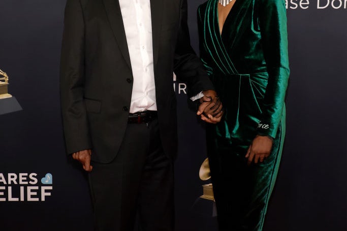 Two people on a red carpet. The person on the left wears sunglasses, a dark suit, and a hat. The person on the right wears a velvet gown