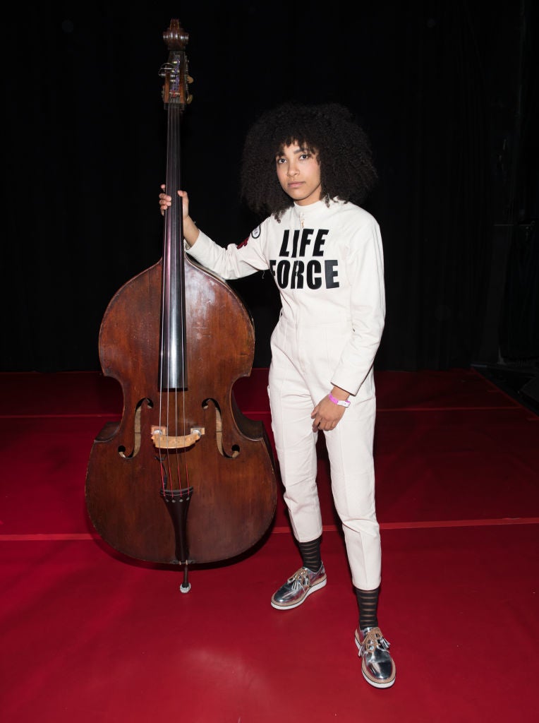 Person in a white jumpsuit with "Life Force" text, standing beside an upright bass on a red carpet