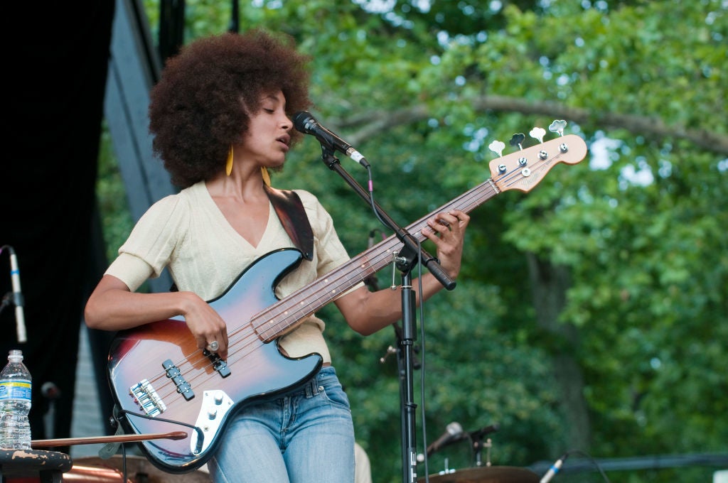Musician playing guitar on stage, wearing a casual light top and jeans, with large earrings, singing into a microphone