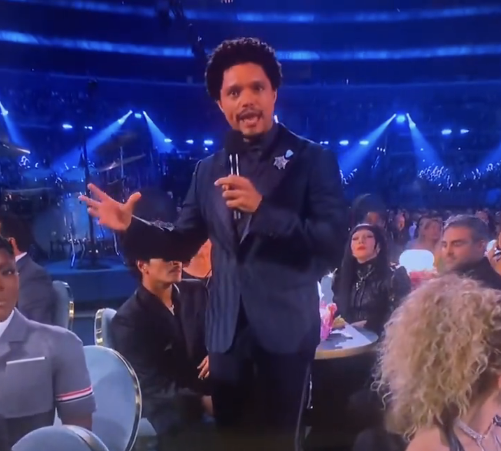 A person in a suit speaks into a microphone at an indoor event with an audience seated at round tables