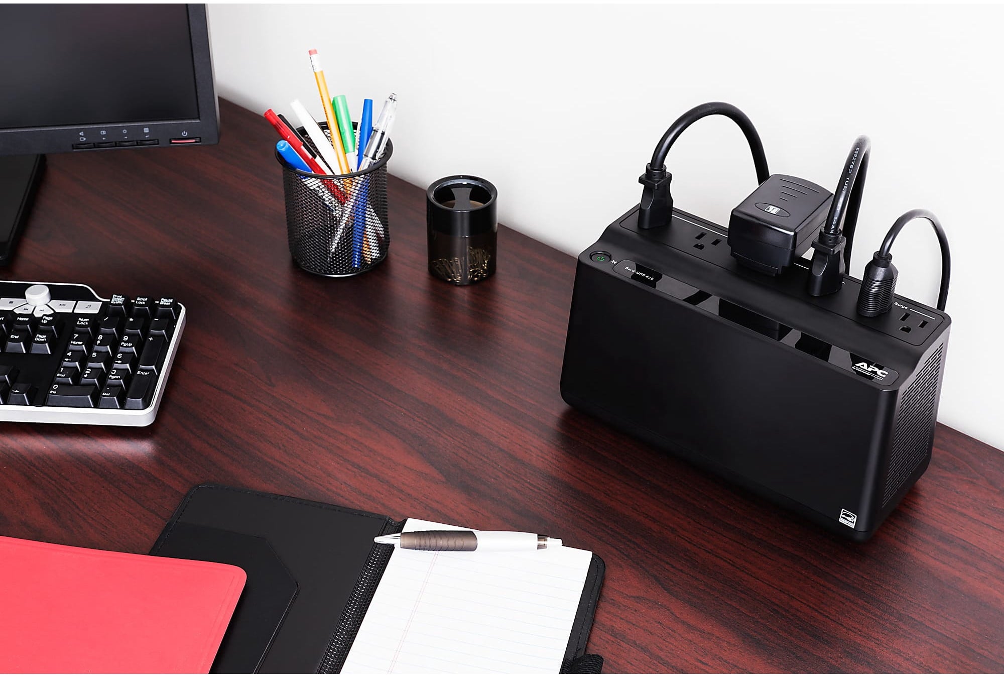 Office desk with a computer keyboard, monitor, pens in a holder, a notebook, and a black UPS device with plugged-in cables