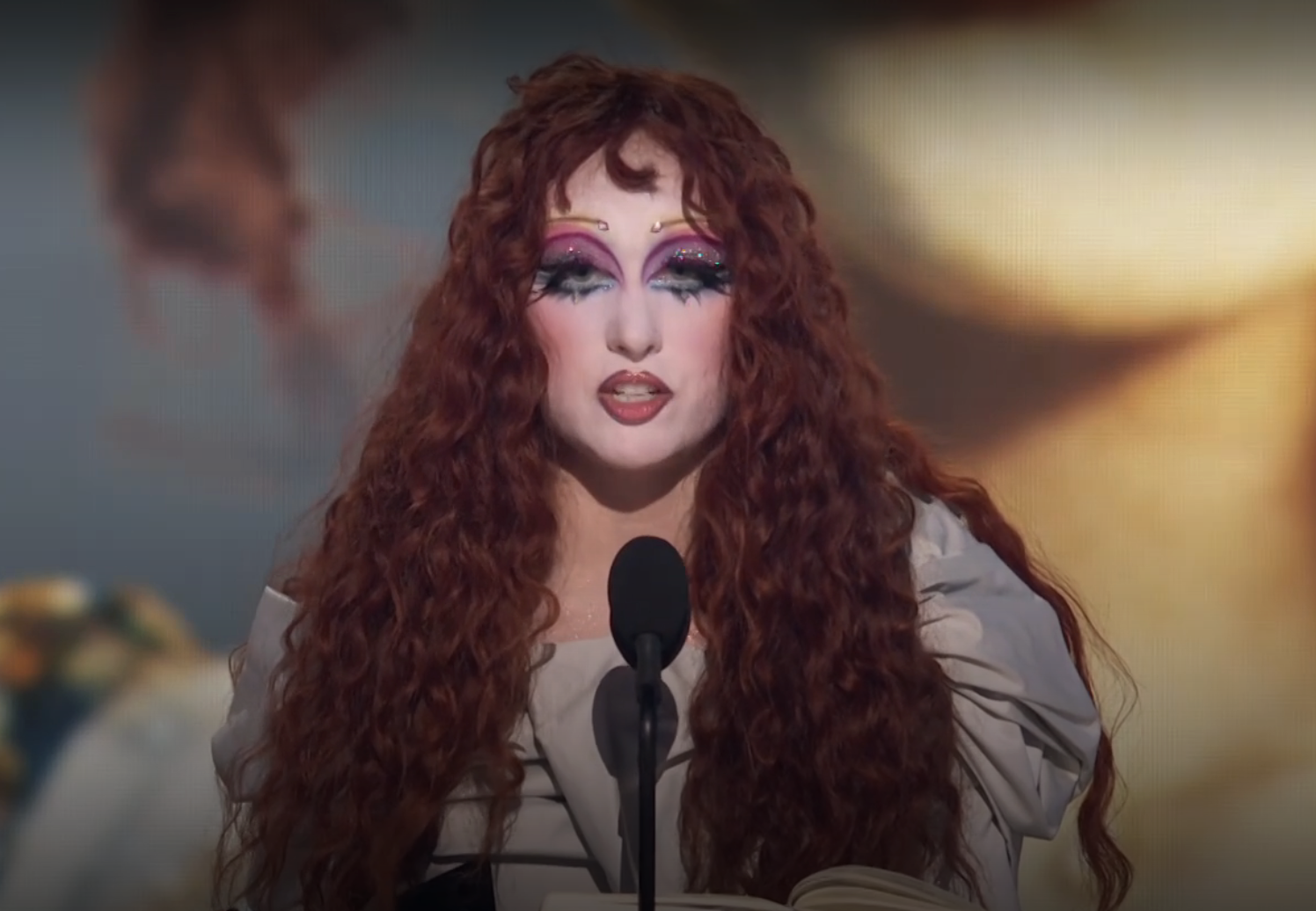 Person with elaborate, dramatic makeup and long, curly hair speaks at a microphone, holding a book