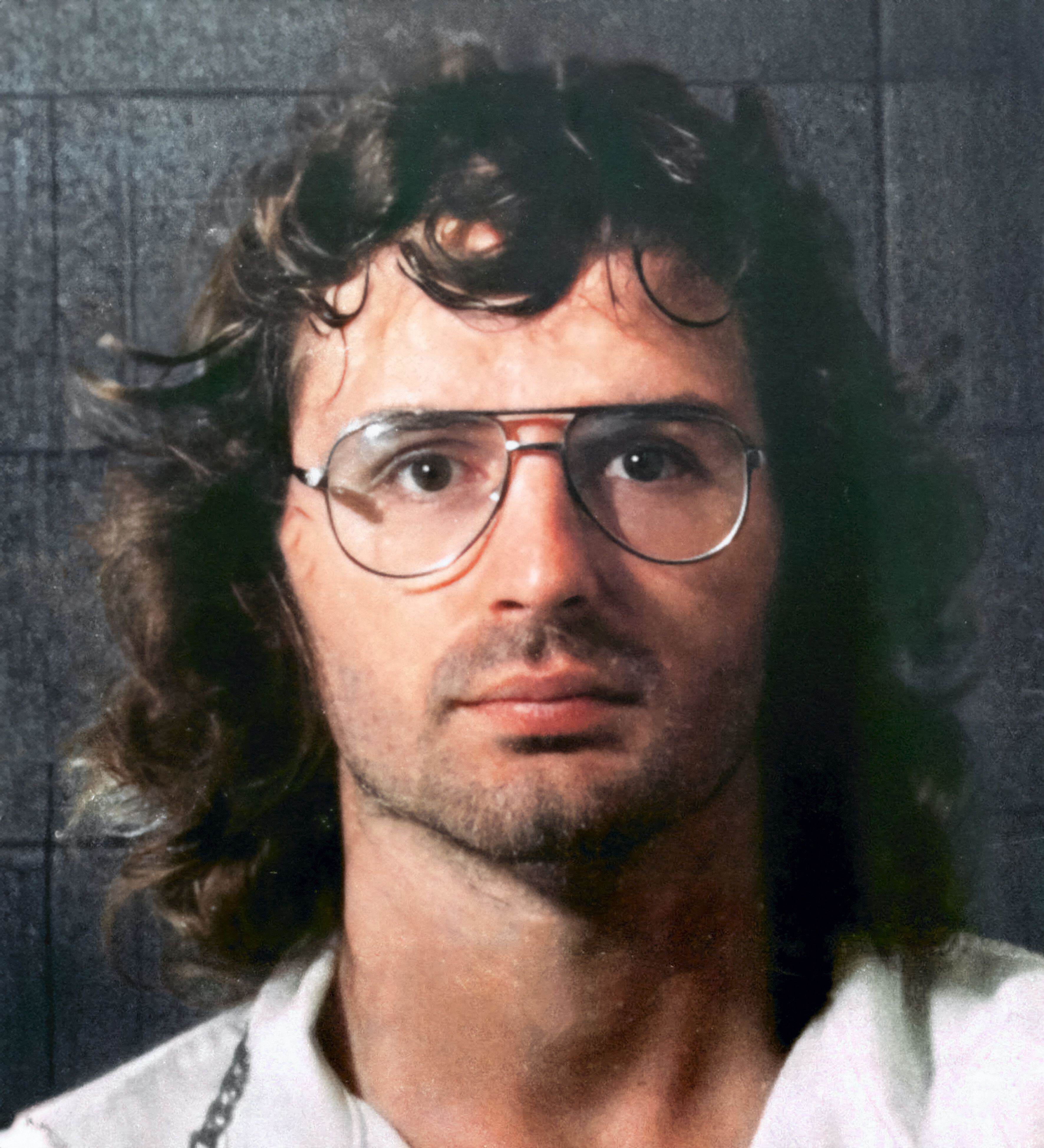 A person with long wavy hair, glasses, and a neutral expression looks directly at the camera