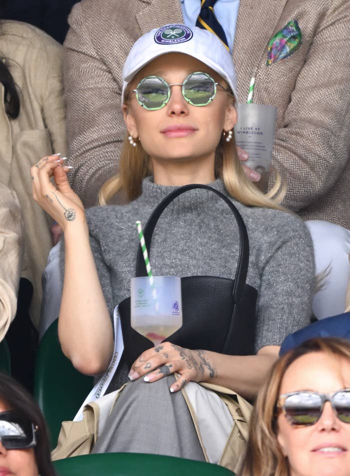 Ariana Grande at a tennis match, wearing a cap, sunglasses, and a gray sweater, holding a drink with a striped straw