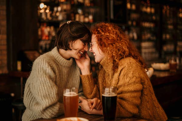 Two people in cozy sweaters lean close, sharing a tender moment at a bar, with drinks in front of them
