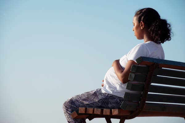 Pregnant woman sitting on a bench, looking thoughtful. She is wearing a white top and patterned pants