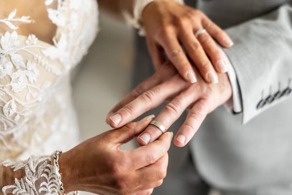 Close-up of a couple exchanging rings, symbolizing marriage or commitment. The bride wears a lace dress, and the groom dons a light suit