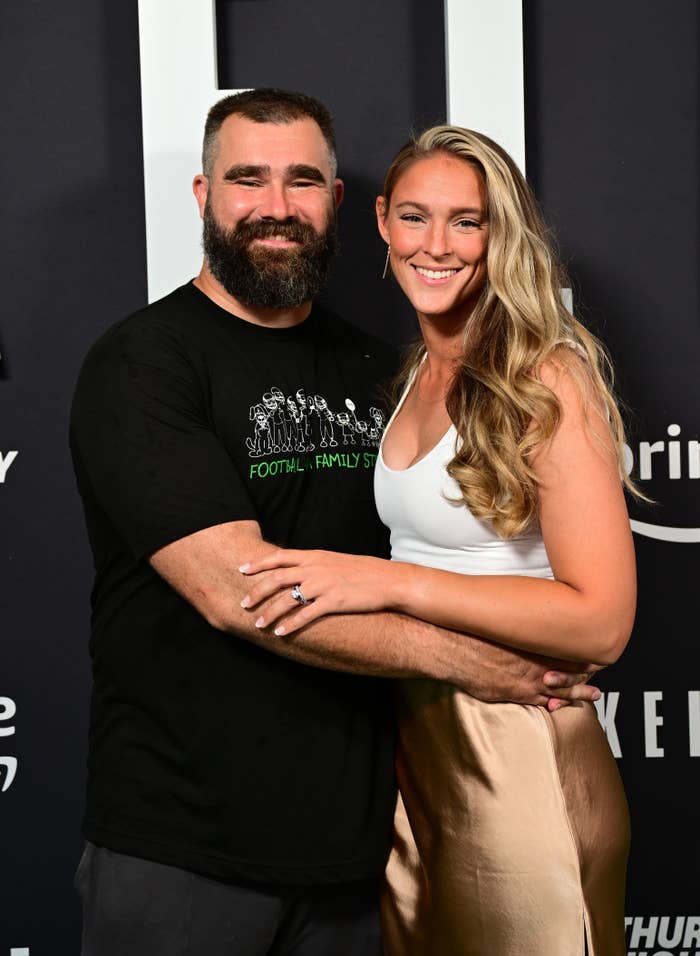 Jason Kelce wears a casual T-shirt and has his arm around Kylie Kelce who is wearing a white top with a satin skirt