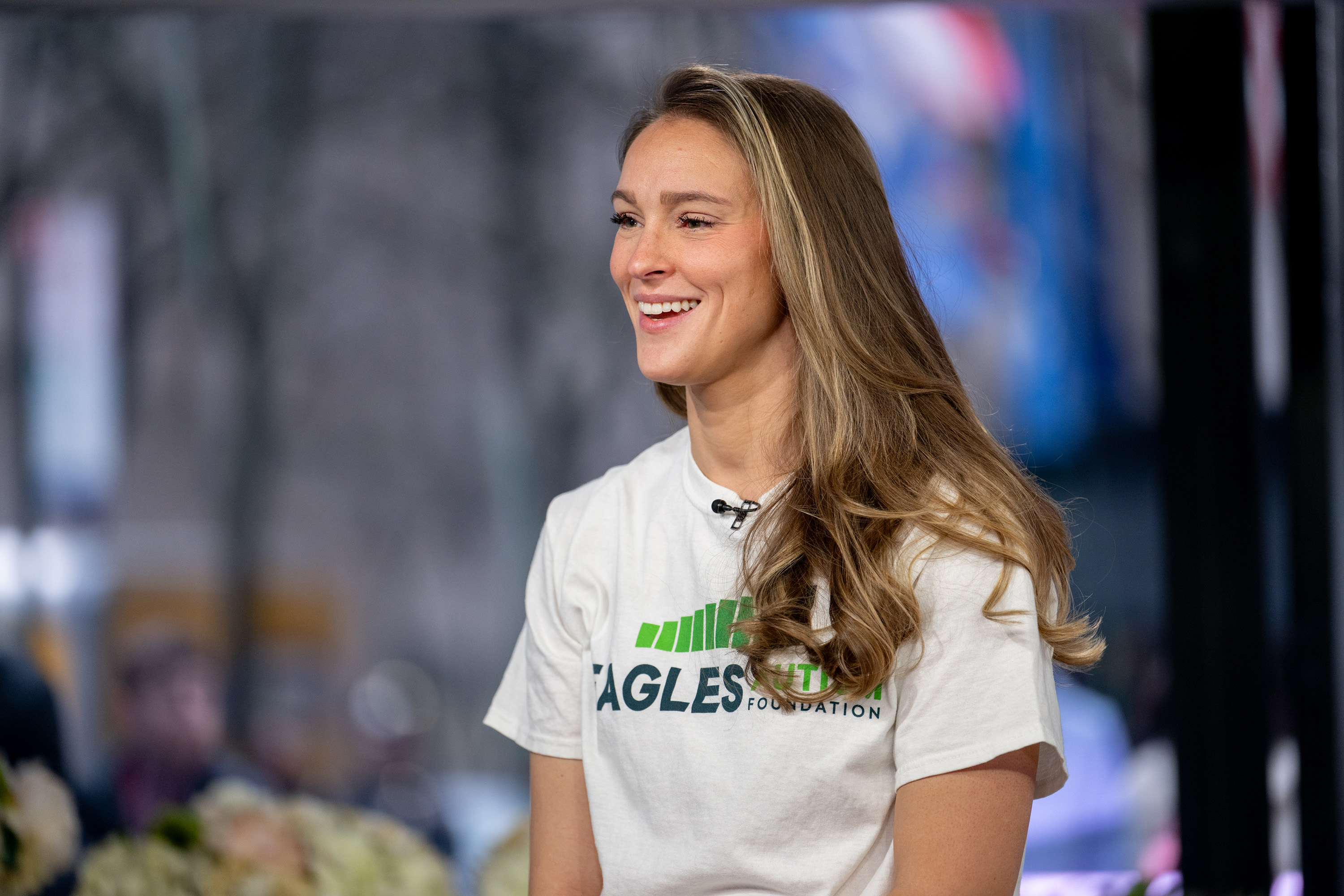 Kylie Kelce smiling, wearing a casual Eagles T-shirt with visible text, seated in an indoor setting with a blurred background