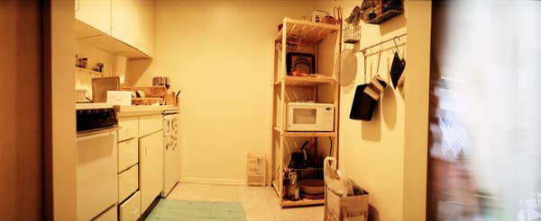 Cozy kitchen with a microwave on a wooden shelf, pots and pans hanging. Simple decor with small appliances and a rug on the floor