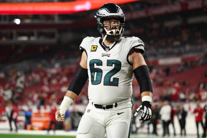 Jason Kelce in football uniform on field, wearing helmet and pads, appears ready for action. Stadium seats in the background