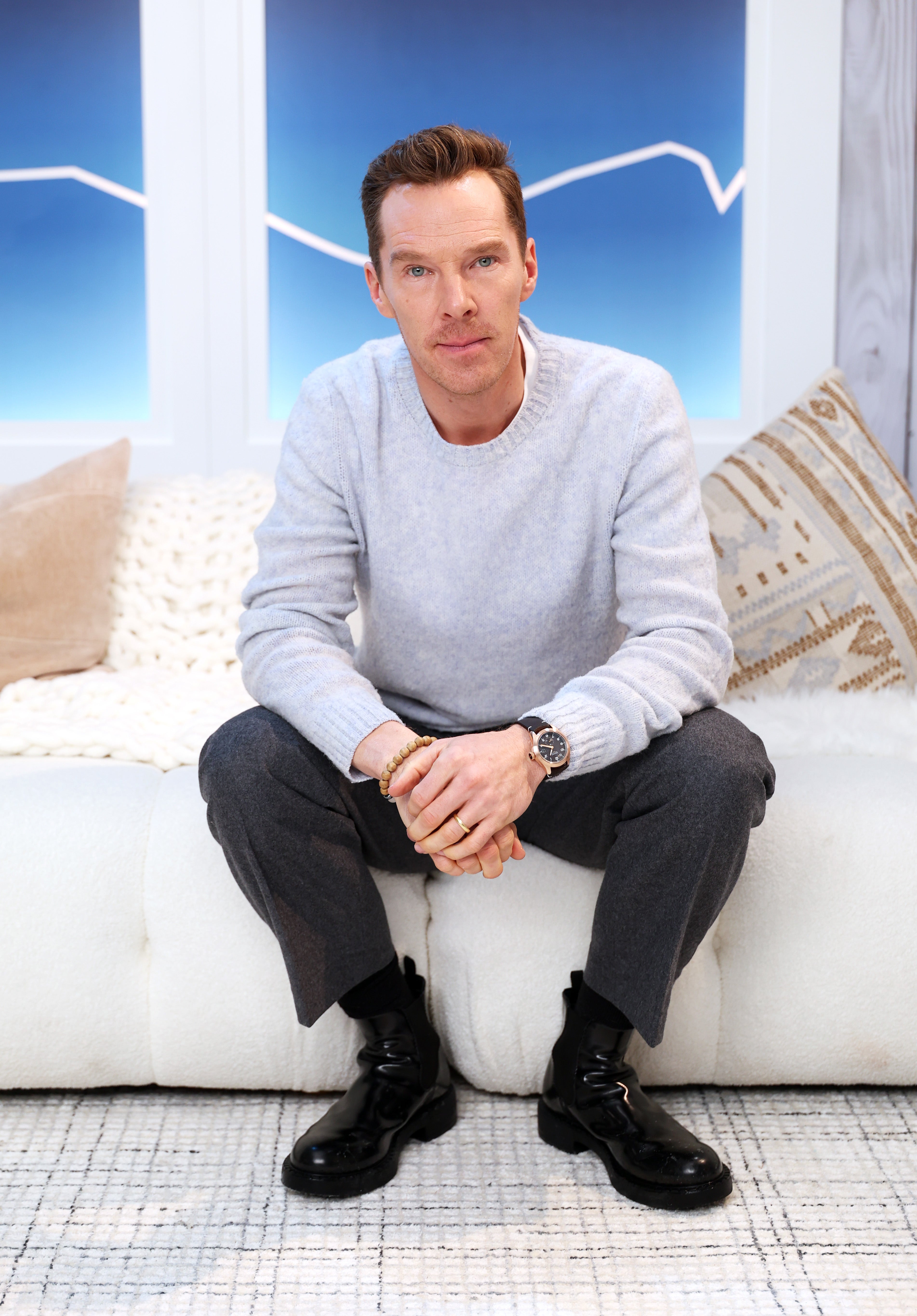 Benedict Cumberbatch sits on a sofa wearing a light sweater, dark trousers, and shiny black shoes, in a modern indoor setting with geometric patterned decor