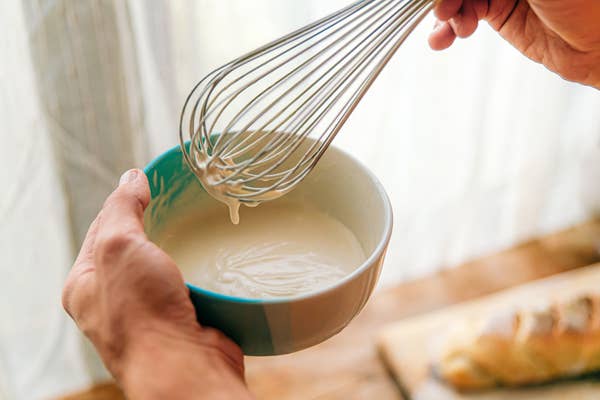 Hands whisking thick batter in a bowl near a window, with bread blurred in the background