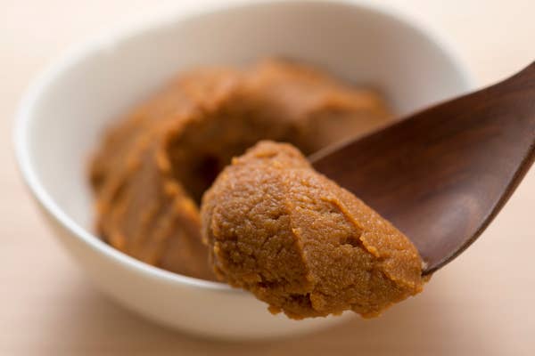 A wooden spoon with a scoop of brown paste, likely miso, over a white bowl containing more of the same paste