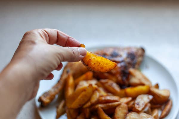 Hand holding a seasoned potato wedge above a plate filled with more wedges and grilled meat