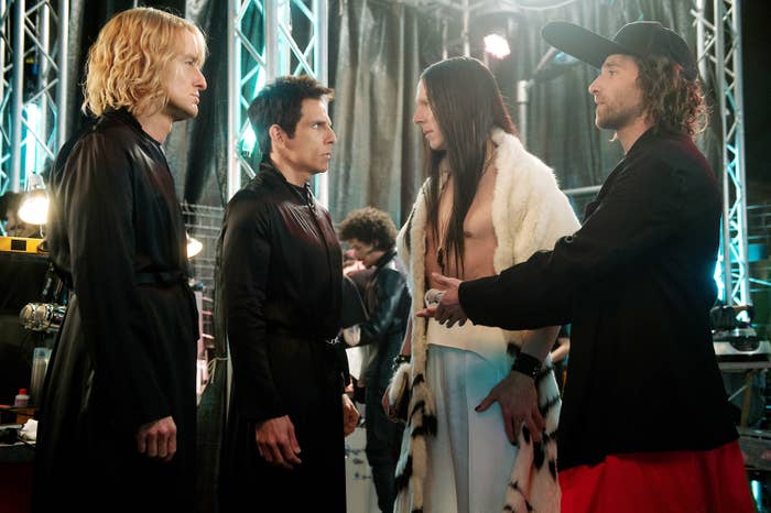 Owen Wilson, Ben Stiller, Benedict Cumberbatch, and Kyle Mooney stand in conversation backstage. Three wear stylish outfits, one has a visible fur coat. They appear engaged in a discussion