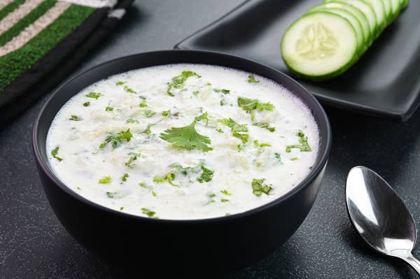 A bowl of yogurt-based cucumber raita garnished with cilantro, next to cucumber slices and a spoon on a table