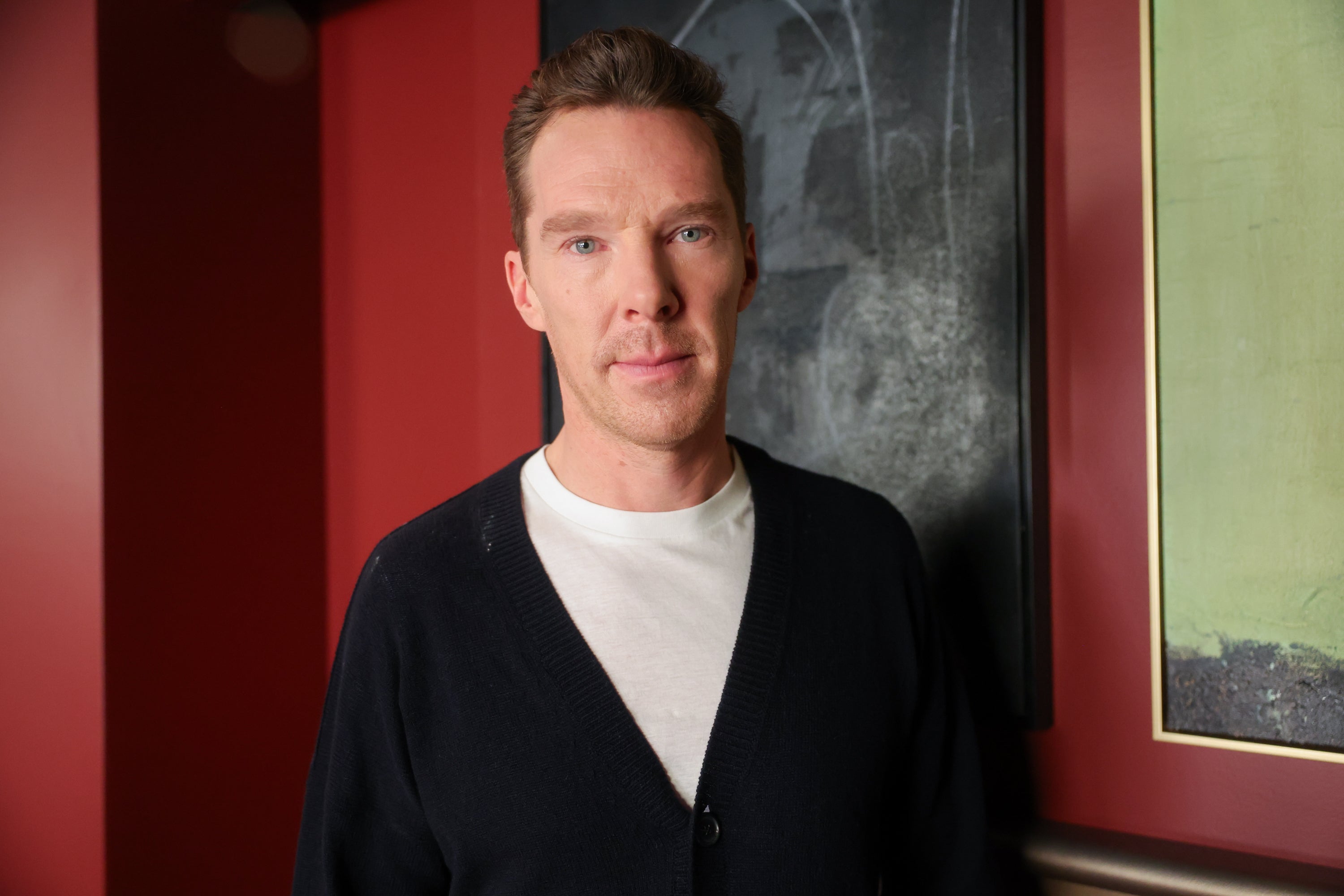 Benedict Cumberbatch in a white shirt and dark cardigan is standing in front of a red wall with framed artwork