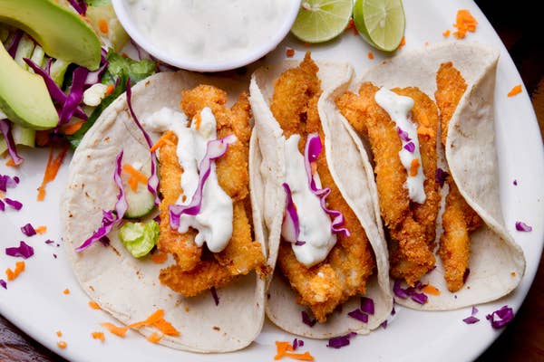 Four crispy fish tacos with shredded cabbage, drizzled with white sauce, served with lime and avocado slices on a white plate