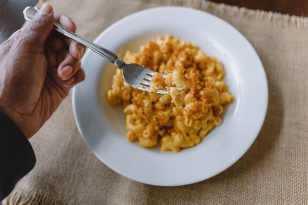 A hand holding a fork with a bite of macaroni and cheese over a plate on a burlap table setting