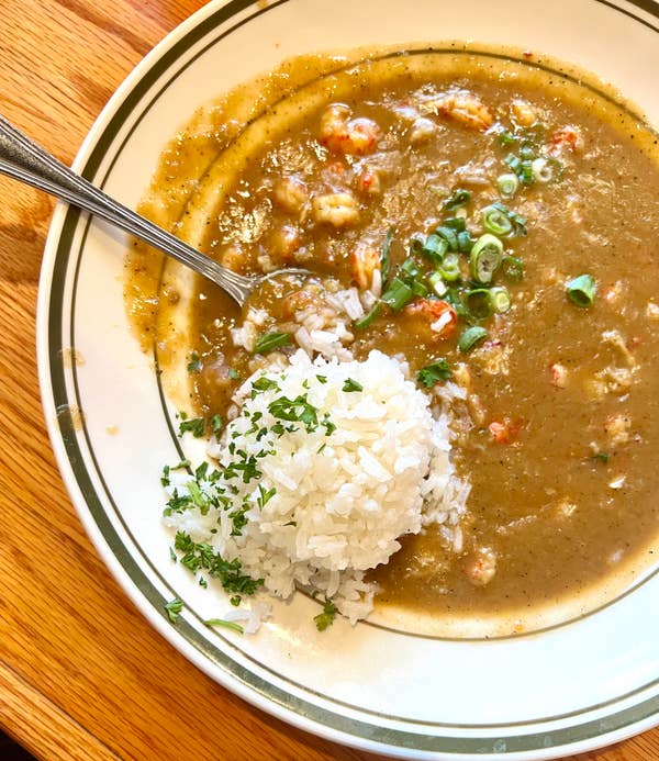 A bowl of gumbo with shrimp and green onions, served with a side of white rice garnished with herbs on a plate with a spoon