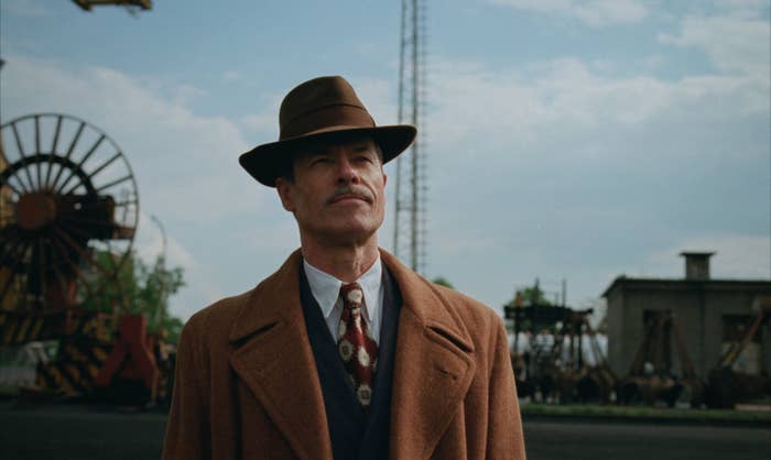 Guy Pearce in a vintage suit and tie with a fedora stands outdoors near industrial structures