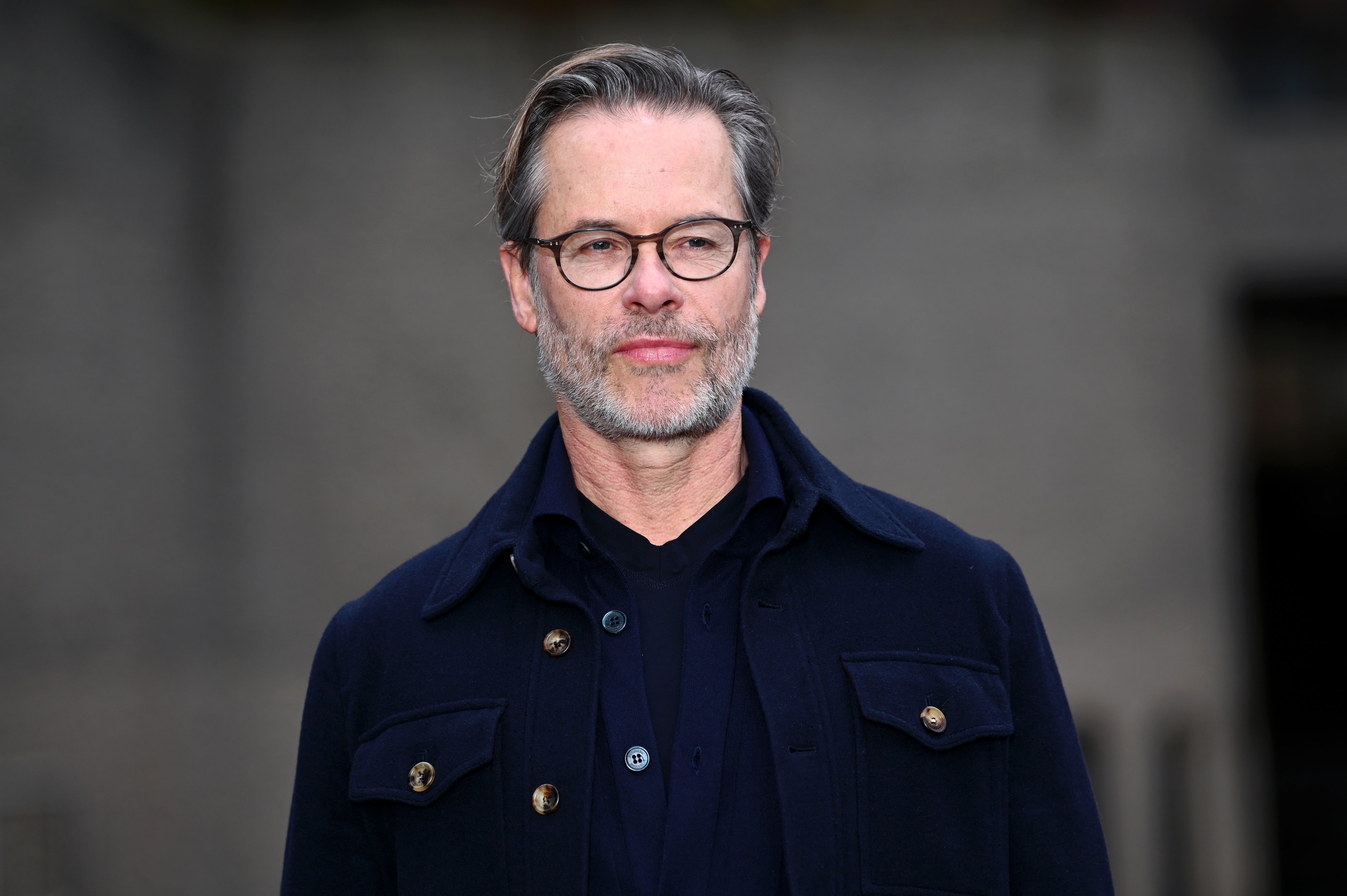 Guy Pearce with glasses, wearing a dark jacket over a shirt, stands outdoors