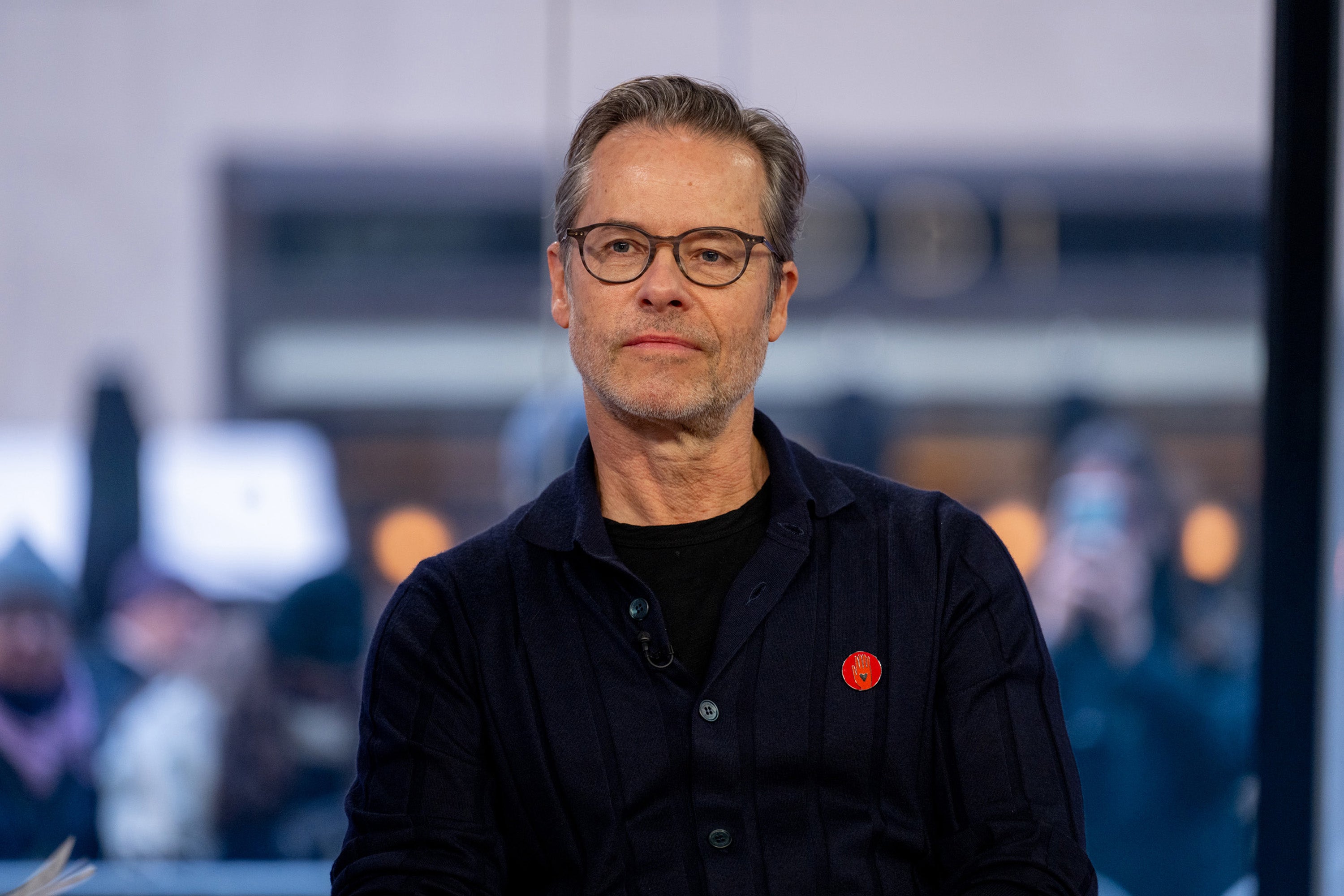 Guy Pearce with glasses wearing a dark jacket and a red emblem, seated indoors during an interview or discussion, with a blurred background