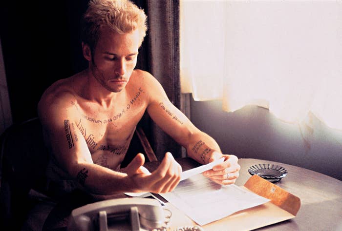 Guy Pearce sits at a table, examining a paper. He has multiple tattoos on his arms and chest
