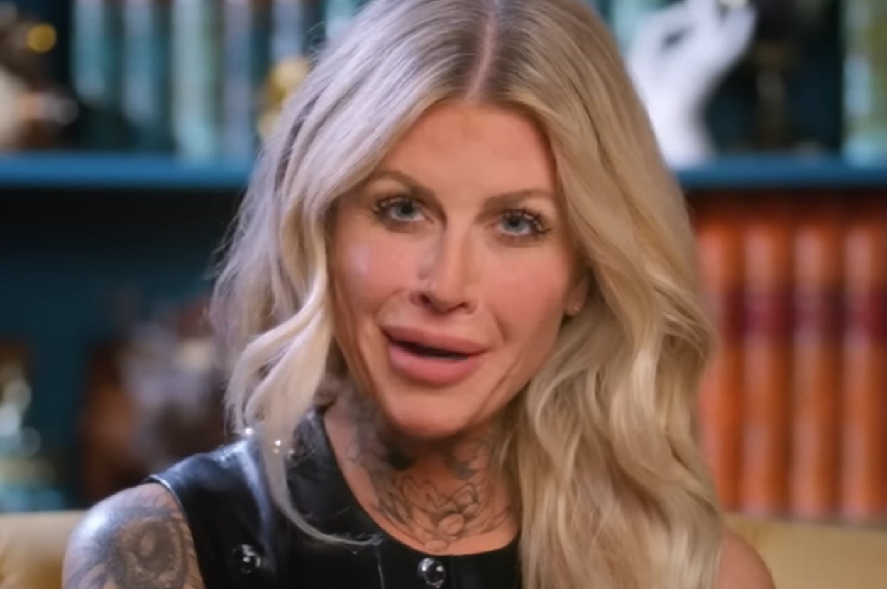 A person with long wavy hair and visible tattoos is smiling and wearing a sleeveless, shiny top. Books are blurred in the background
