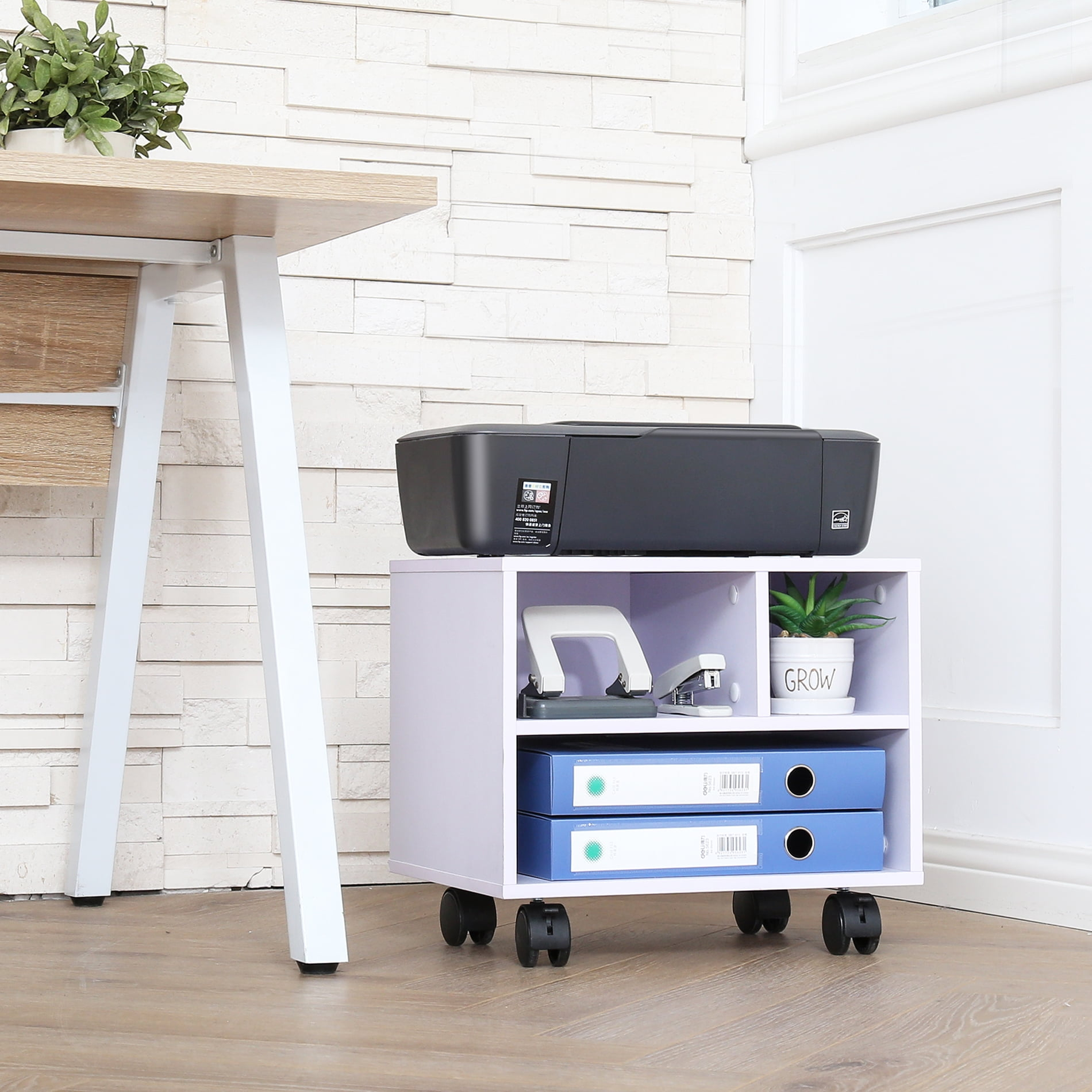 Printer on a white rolling shelf with stationery, binders, and a plant, placed under a desk against a white brick wall