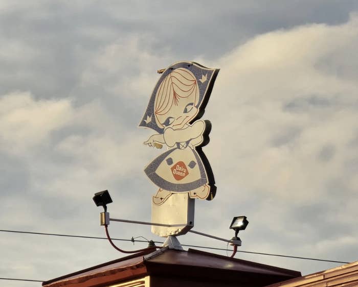 Sign of a cartoon character dressed as a dough girl atop a building, with "Dixie Queen" on the dress. Cloudy sky in the background