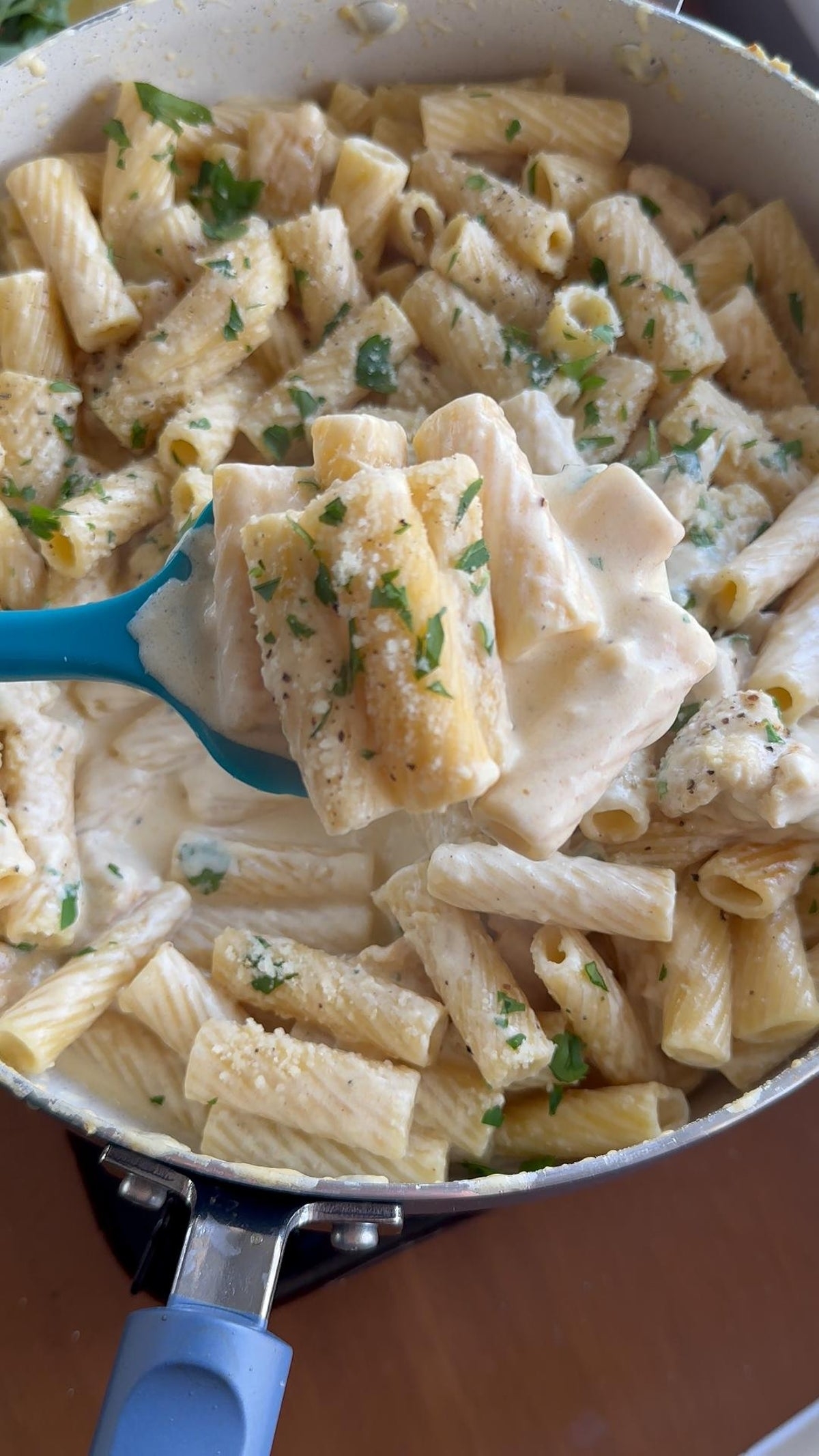 Creamy rigatoni pasta in a pan, garnished with herbs and grated cheese, being served with a blue spoon