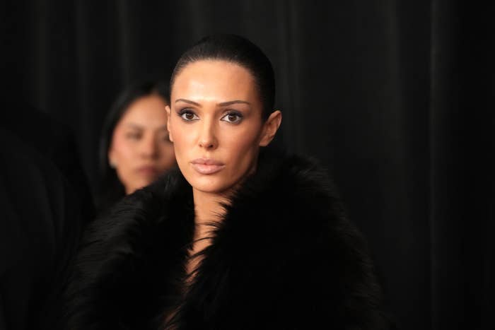 Person in elegant fur-trimmed outfit, with slicked-back hair, stands against a dark backdrop, focused expression