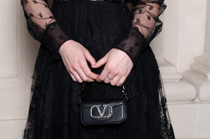 A celebrity poses in a sheer lace dress with a black skirt and matching heels, holding a small clutch, at a formal event indoors