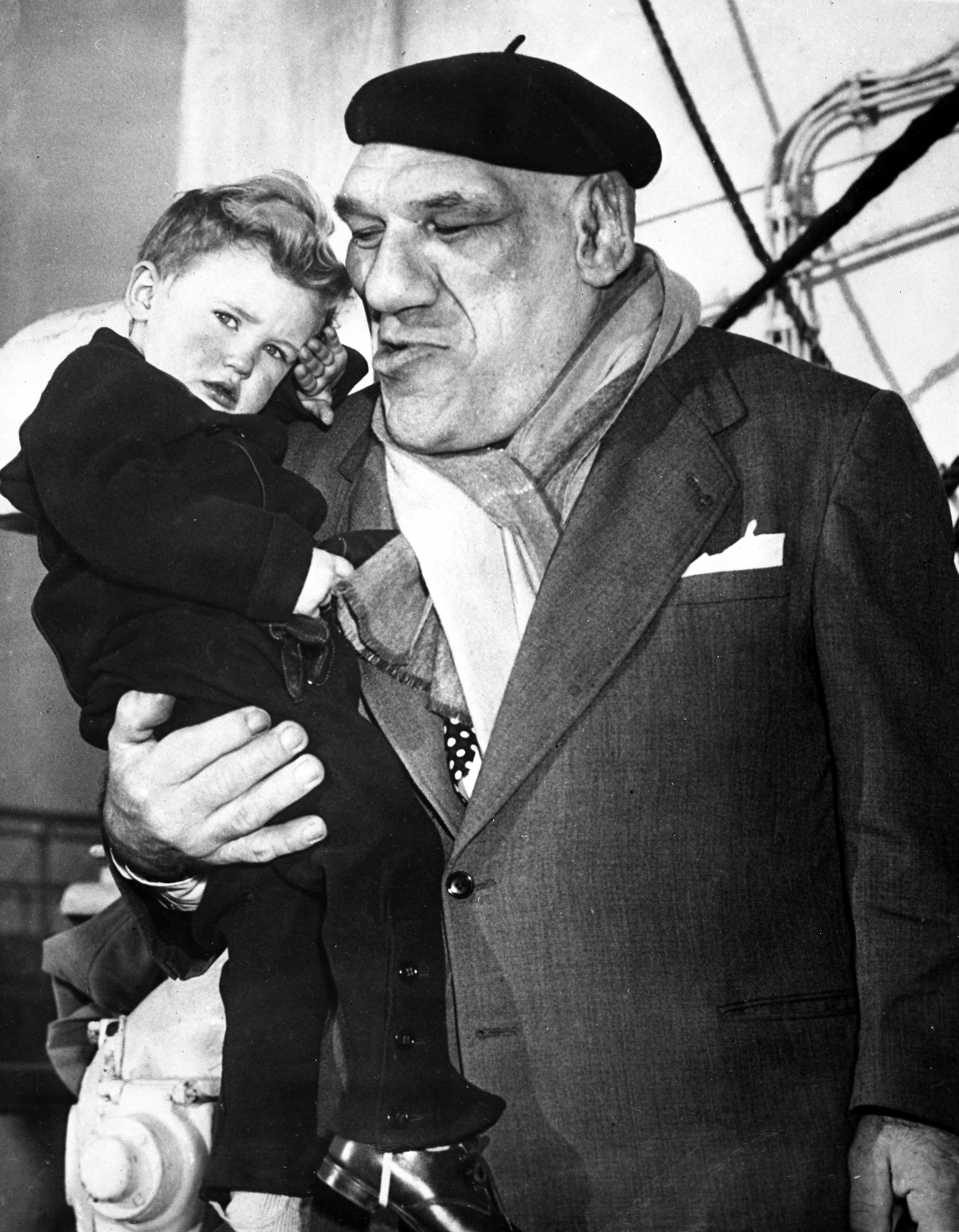 An older man in a suit, scarf, and beret holds a young child, who has a serious expression, aboard a ship