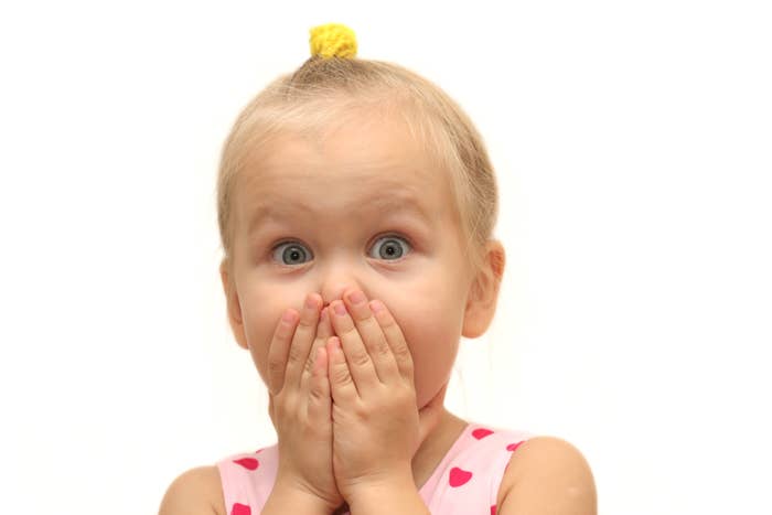 Child with a surprised expression, hands covering mouth, wearing a polka dot dress, hair in a topknot