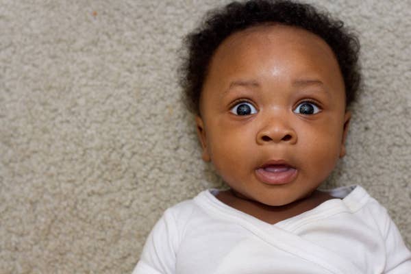 Baby lying on carpet, looking up with a surprised expression, wearing a simple onesie