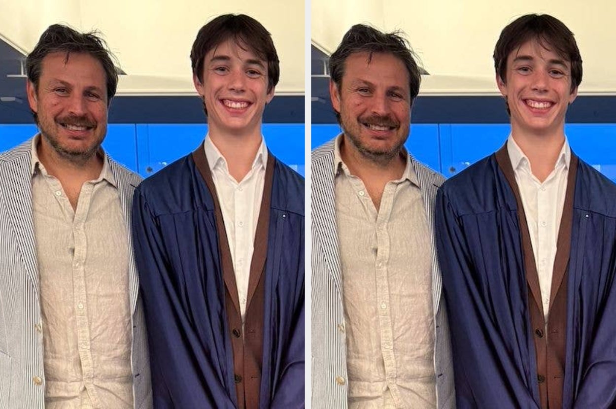 A young graduate in a gown and a striped blazer stands happily with an adult in a casual blazer and shirt, both smiling for a photo