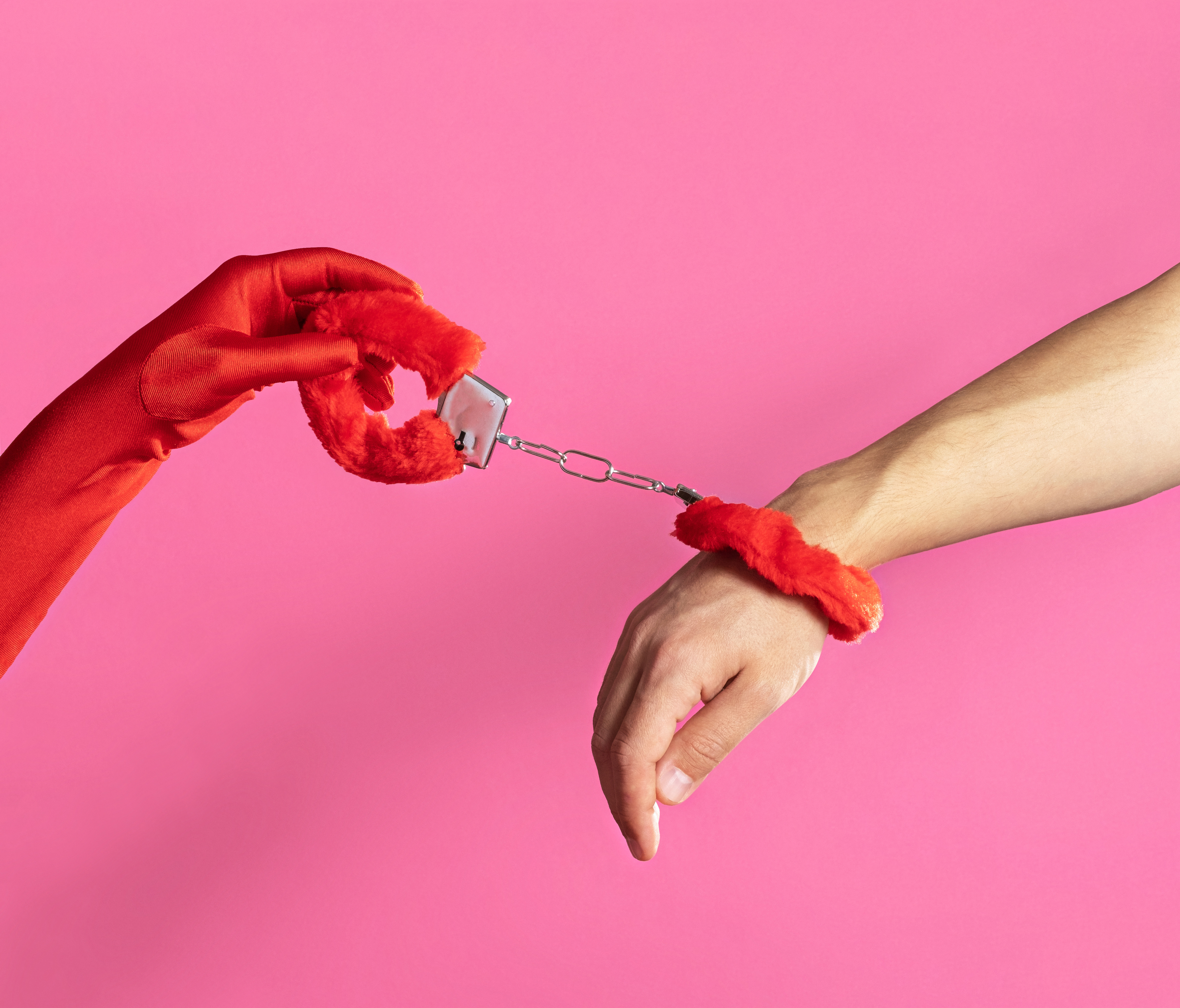 A gloved hand holds a furry red handcuff. The other handcuff is attached to a man's wrist