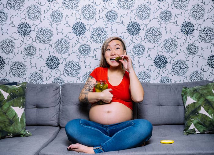 Pregnant woman in casual wear sits on a couch, eating pickles, with a floral-patterned wall in the background