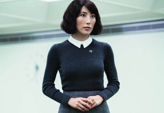 Person in a formal outfit with a collared sweater and skirt, standing in an indoor setting, engaged in conversation