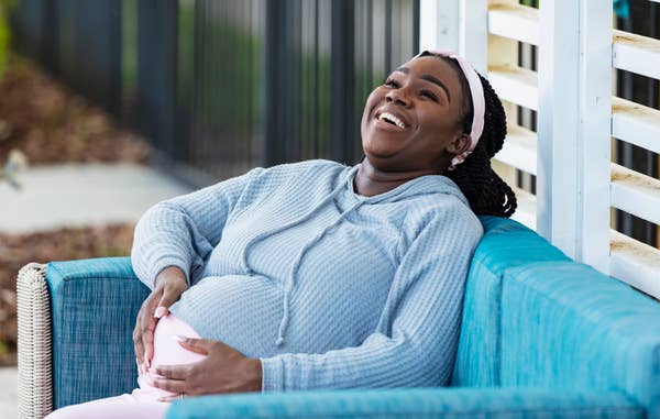 Pregnant person smiling, sitting on a couch outdoors, wearing a comfortable sweater and headband, hands resting on their belly