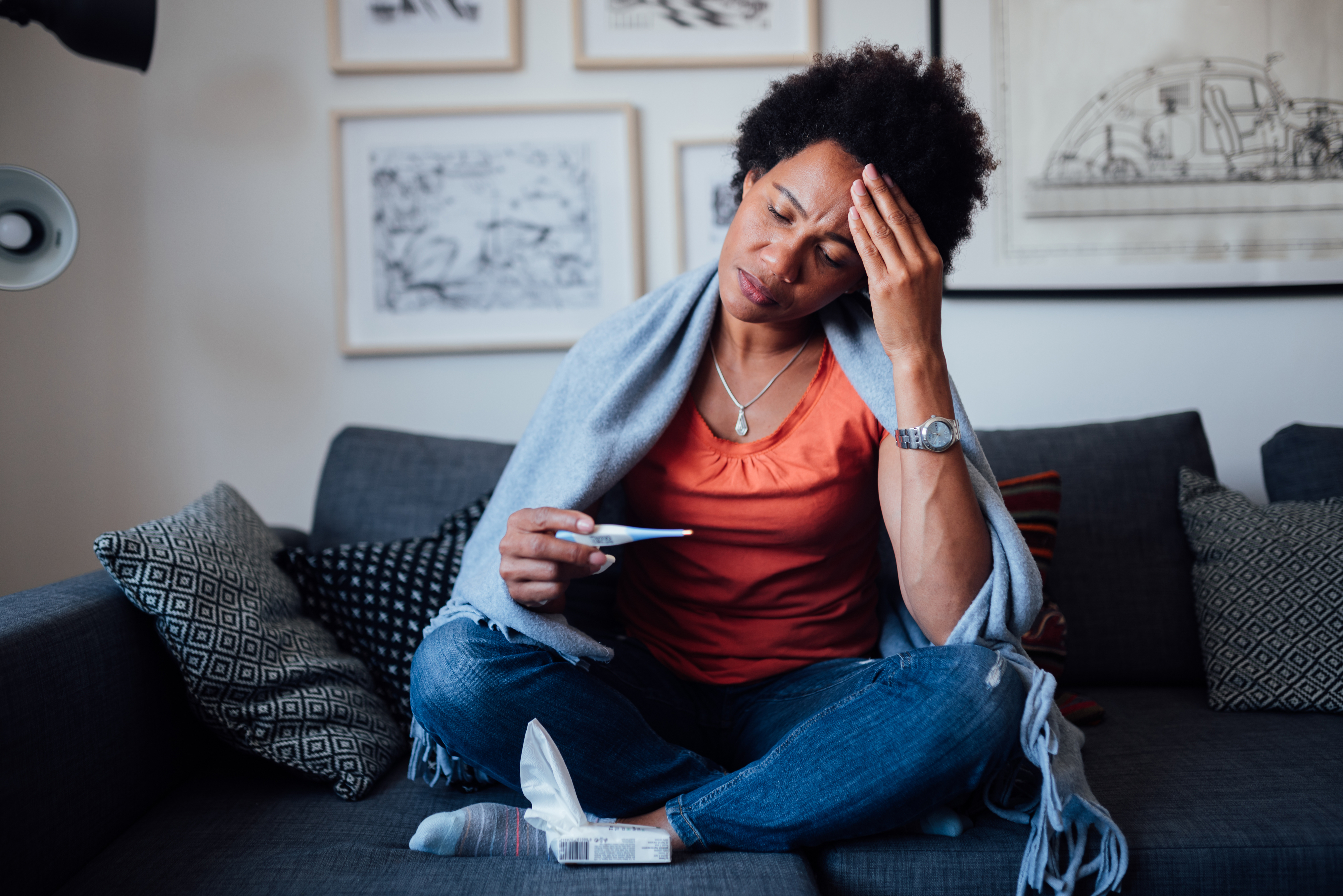 Personen som sitter på en sofa, ser på et termometer med en hånd på hodet, vises uvel, pakket inn i et teppe. Uformell hjemmeinnstilling